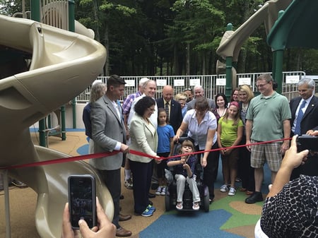 group of colleagues and parents watching a young special needs girl in wheelchair cutting a ribbon at a grand opening at a park