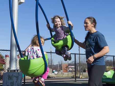 brunette haired teacher pushing two young blonde girl babies smiling in a joint lime green swing with blue handles