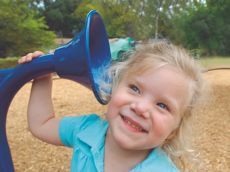 small blonde girl smiling putting her ear up to a blue play speaker phone at a playground