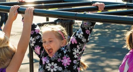 young girl in a flower shirt smiling holding on monkey bars