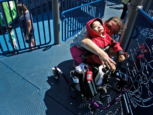 child in wheelchair in red hoodie playing at park with caregiver