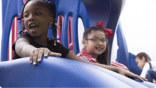 one small african american girl and latina girl with red bow tie in her hair smiling going down a blue slide