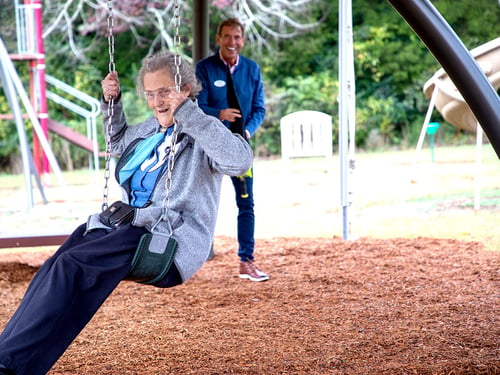 elderly woman swinging at playground with man in blue shirt in the background smiling