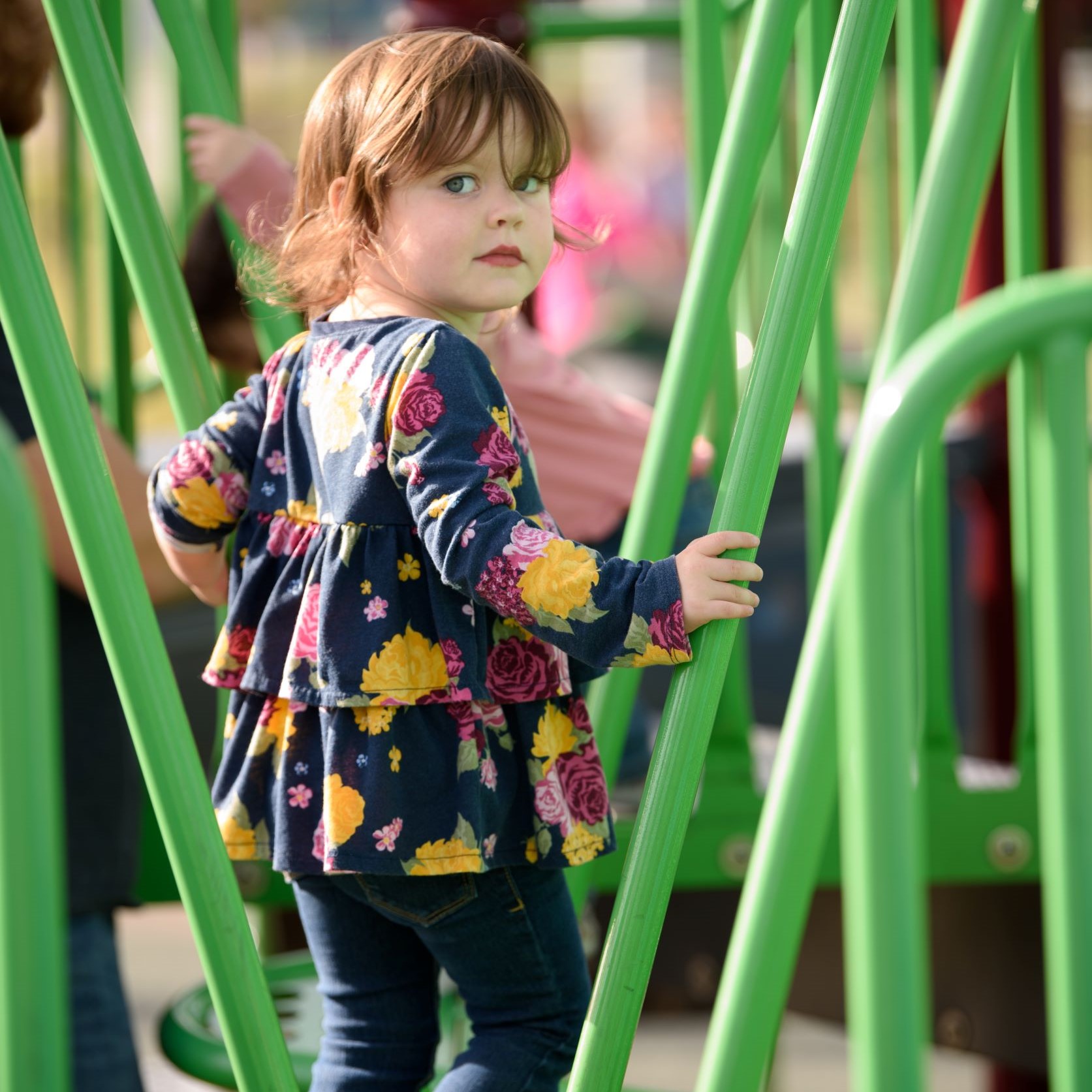 Young girl in flower shirt staring back at the camera as she walks through green playground bars