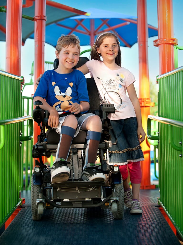 one child in wheelchair with friend having arm around his should on playground set