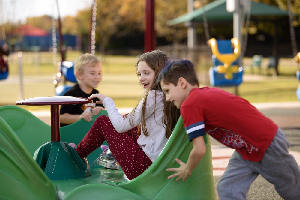 young boy in a red shirt pushing young girl on small green playground equipment