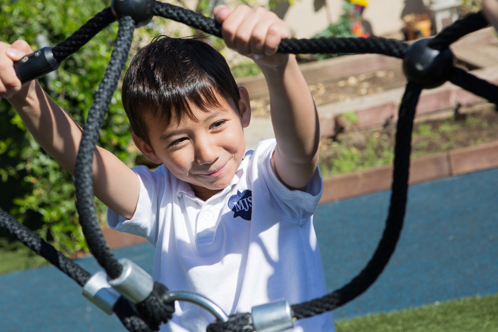young boy with black hair in white shirt smiling holding on black rope at park