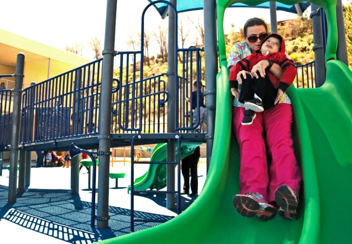 mom holding son in red jacket going down green slide