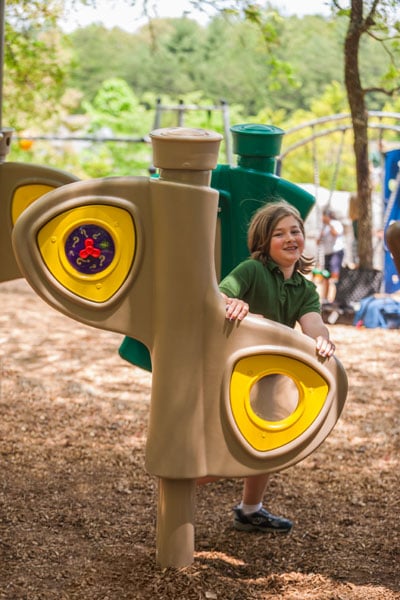 Child in green shirt standing and smiling at the camera while holding onto a brown and yellow playground freestanding structure