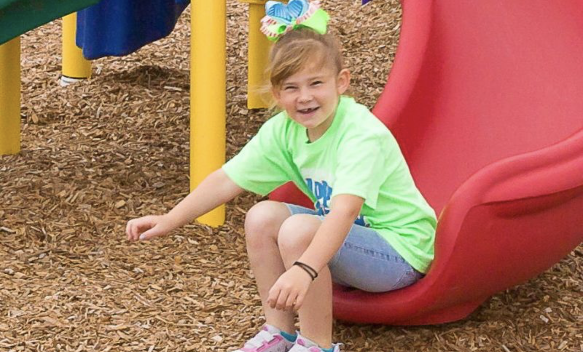 A young girl in bright green shirt smiling at the camera as she sits at the bottom of a red slide