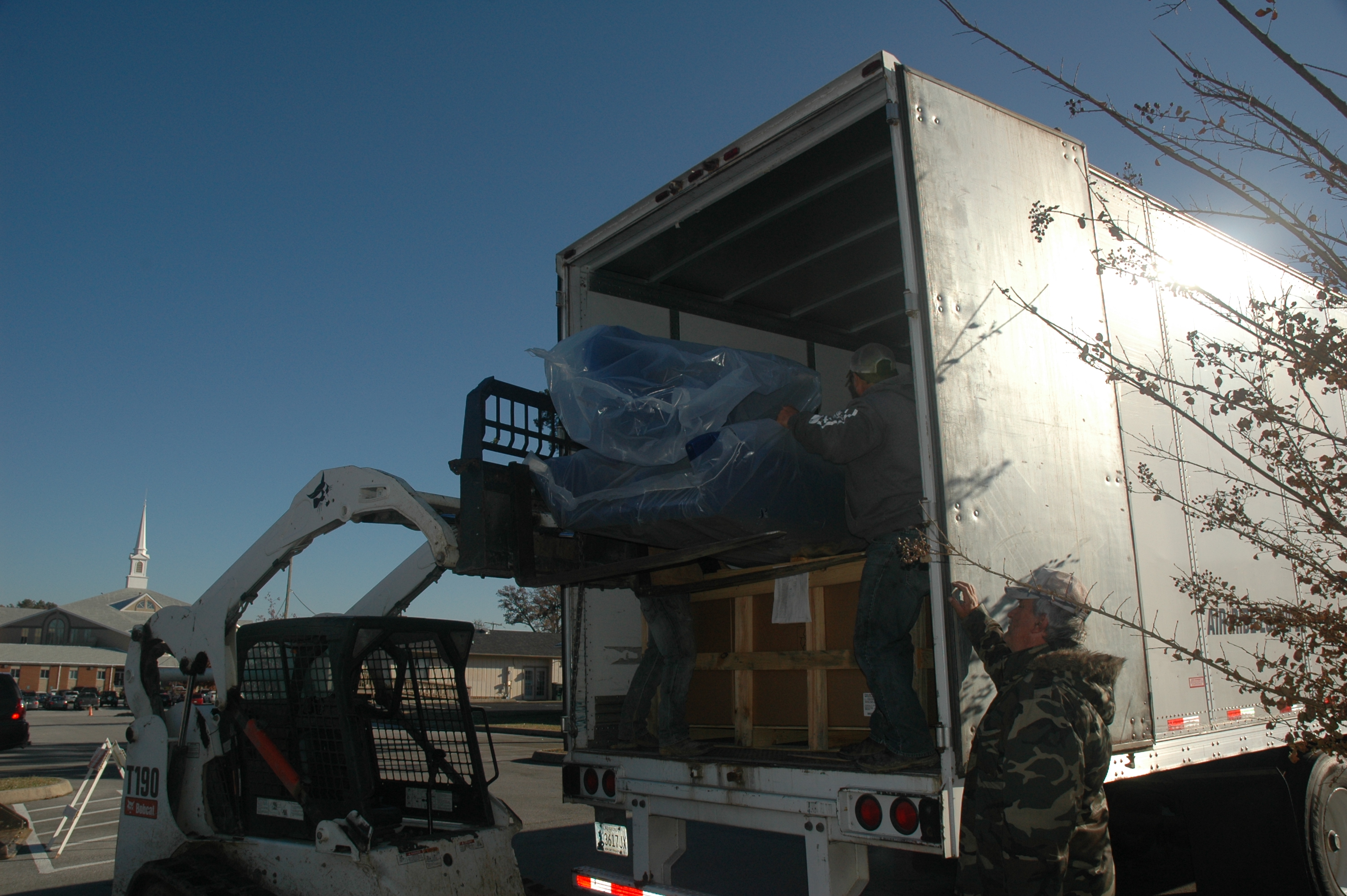 A Bobcat vehicle lowering playground equipment out of the back of a large truck as a man watches